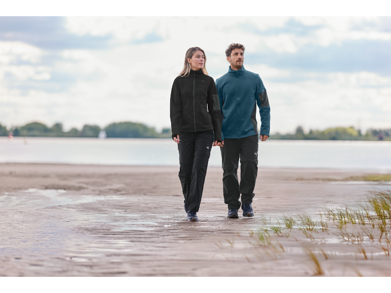 Två personer i fleecejackor och vandringsbyxor går på en strand.