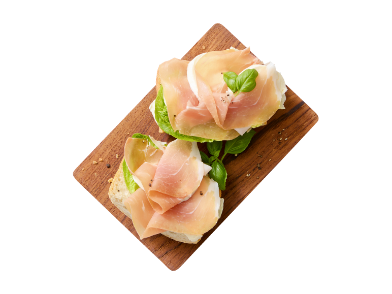 Två öppna smörgåsar med prosciutto, mozzarella, sallad och basilika på en träskärbräda.