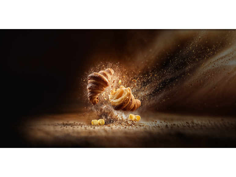 Två gyllene croissanter, en bruten, med smulor och smör som flyger.