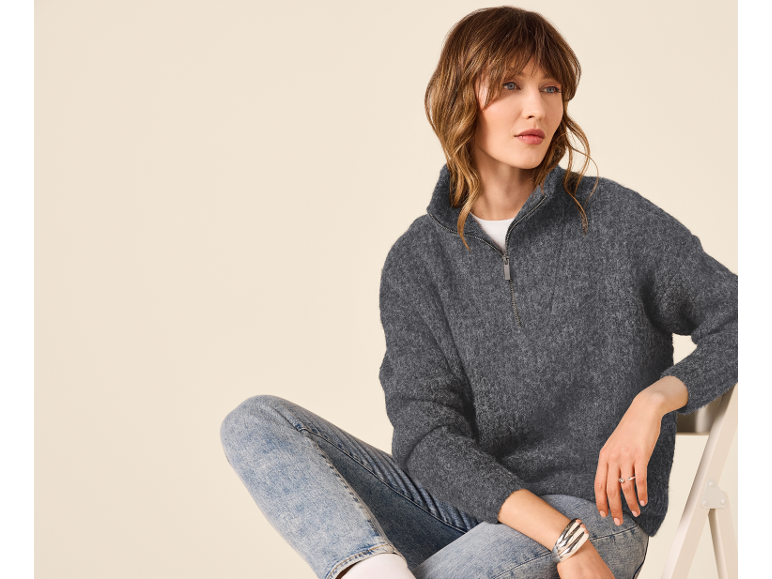 Kvinna i grå dragkedjetröja och jeans, sittande på en stol.