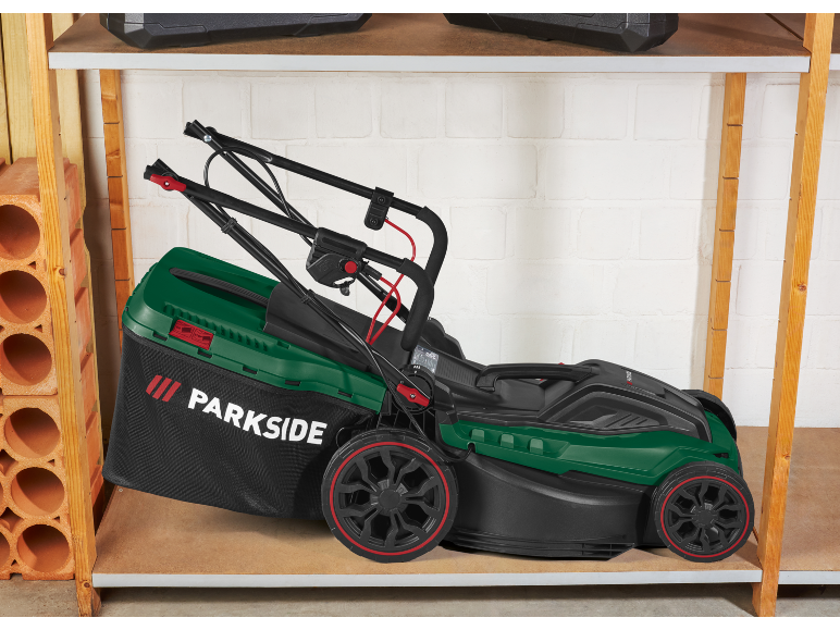 Grön gräsklippare med uppsamlare, förvarad på en trähylla i ett garage.