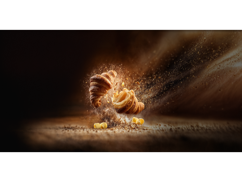 Två gyllene croissanter, en bruten, med smulor och smör som flyger.
