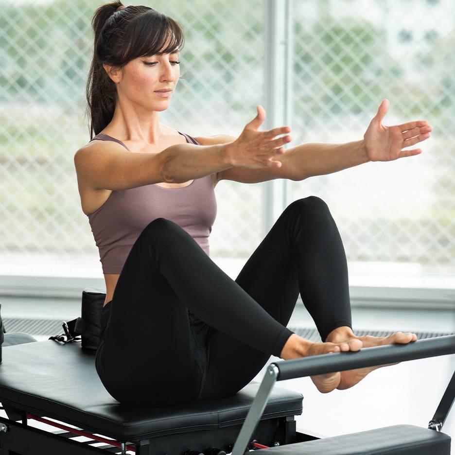 Kvinna som utför Pilates på en reformer, klädd i topp och svarta leggings.