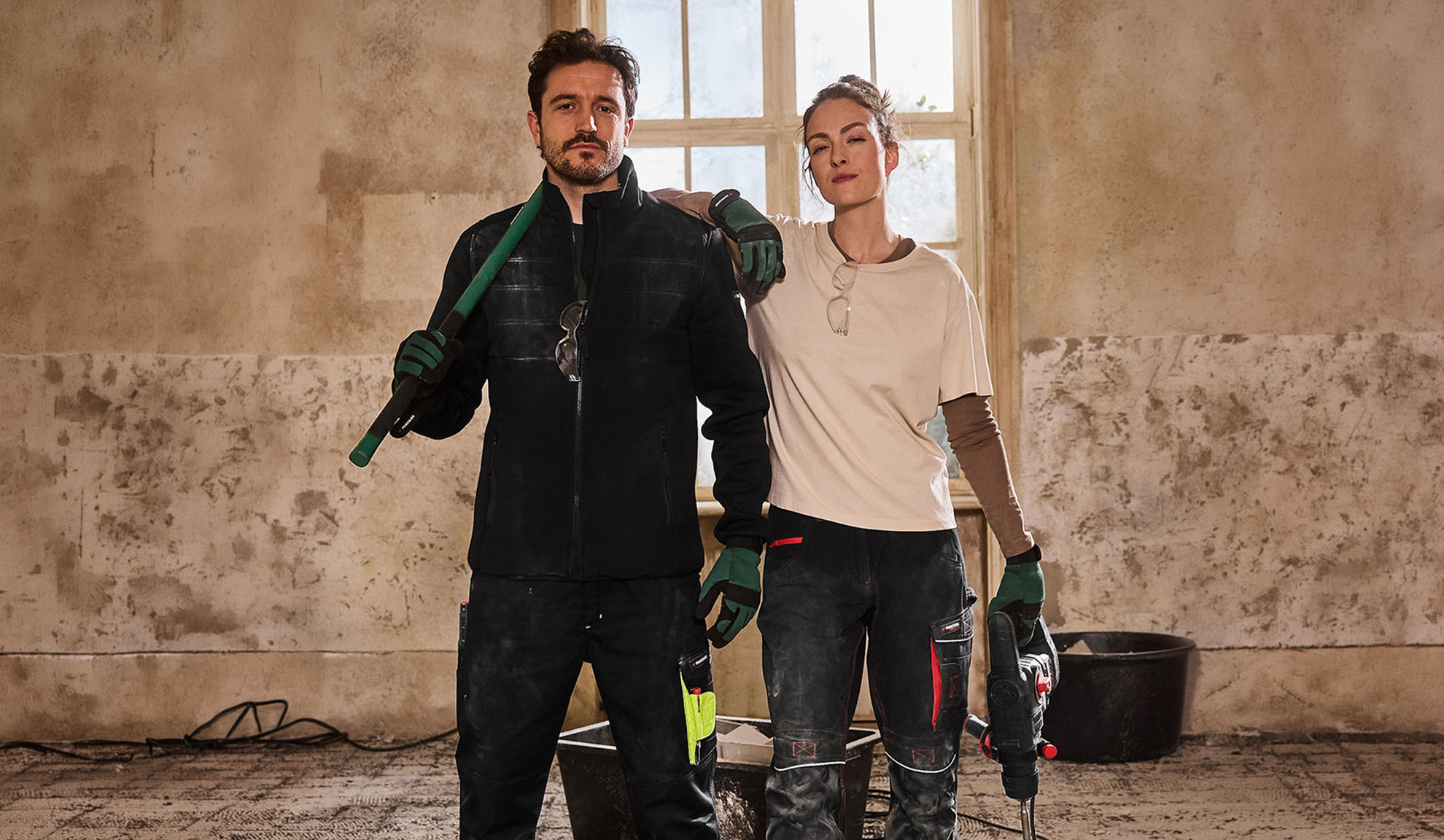 Två byggarbetare i arbetskläder, man med spade och kvinna med borrmaskin.