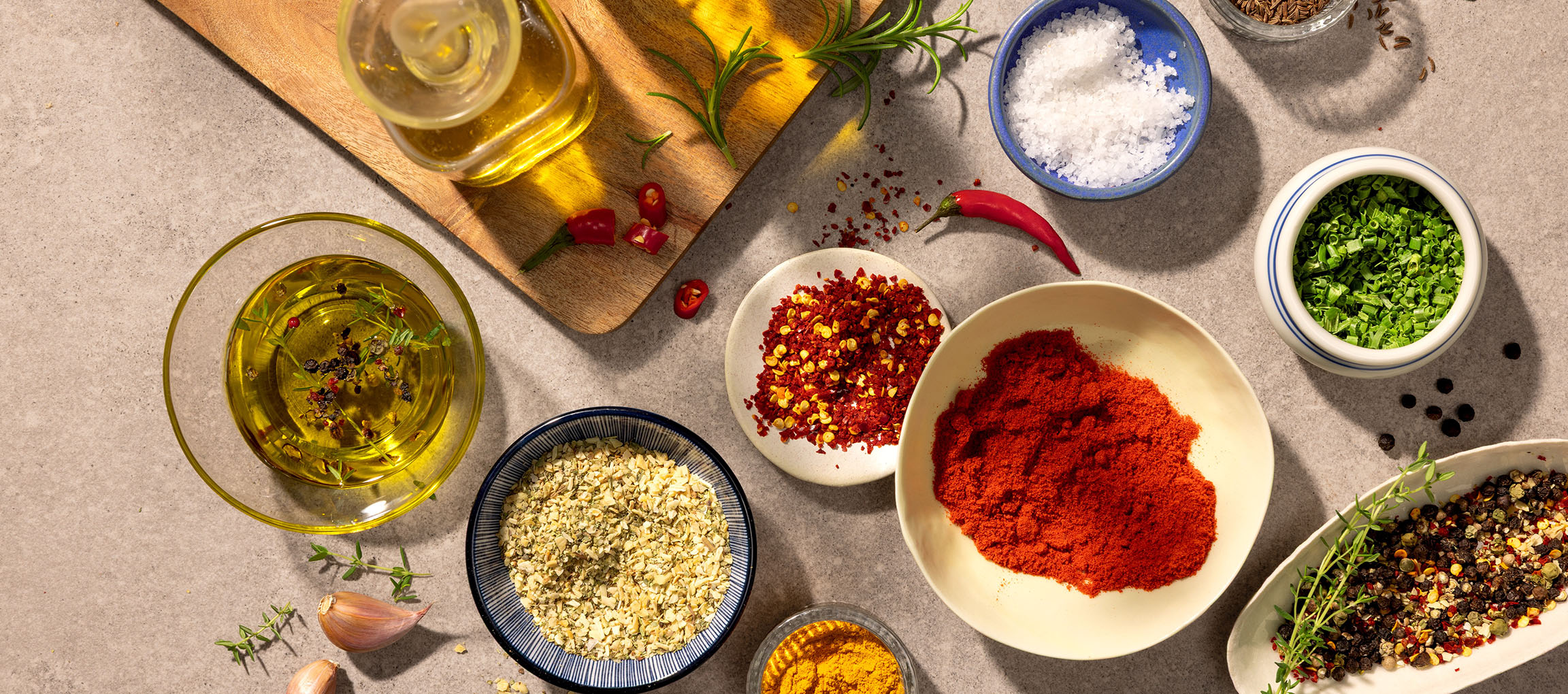 Mångfald av kryddor och oljor, inklusive paprika, chili, salt, peppar och olivolja, på grå bakgrund.