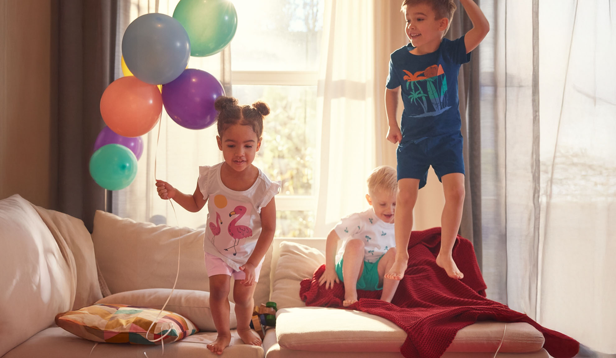 Tre barn hoppar glatt på en soffa med färgglada ballonger.