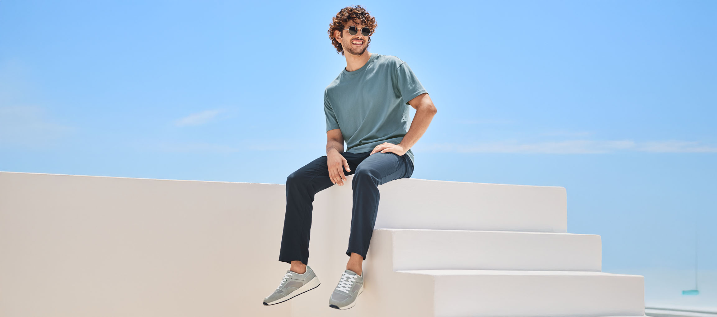 Avslappnad man i t-shirt, byxor och New Balance-skor, sitter på en vit trappa.
