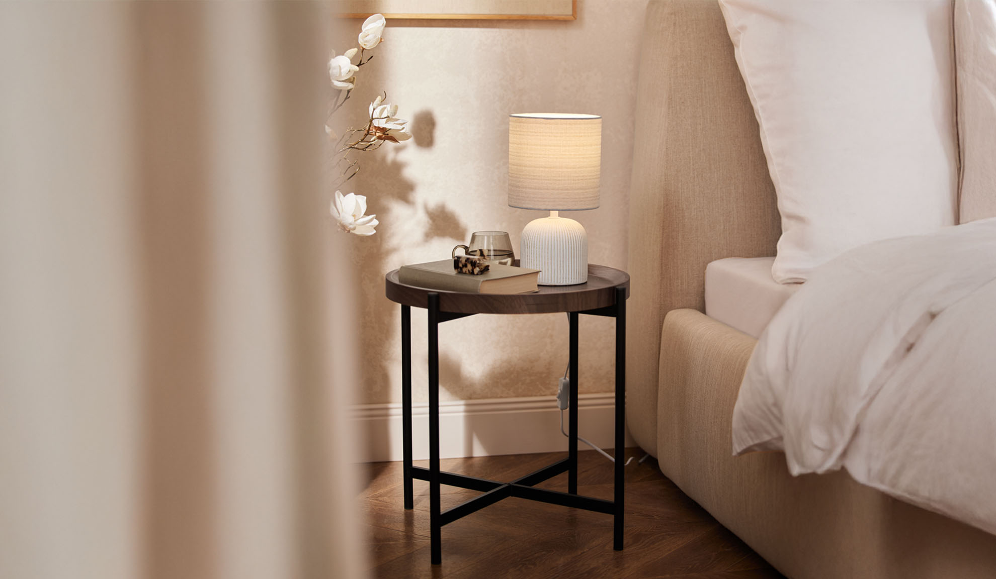 Runt sängbord i trä med bordslampa och tillbehör bredvid en säng.