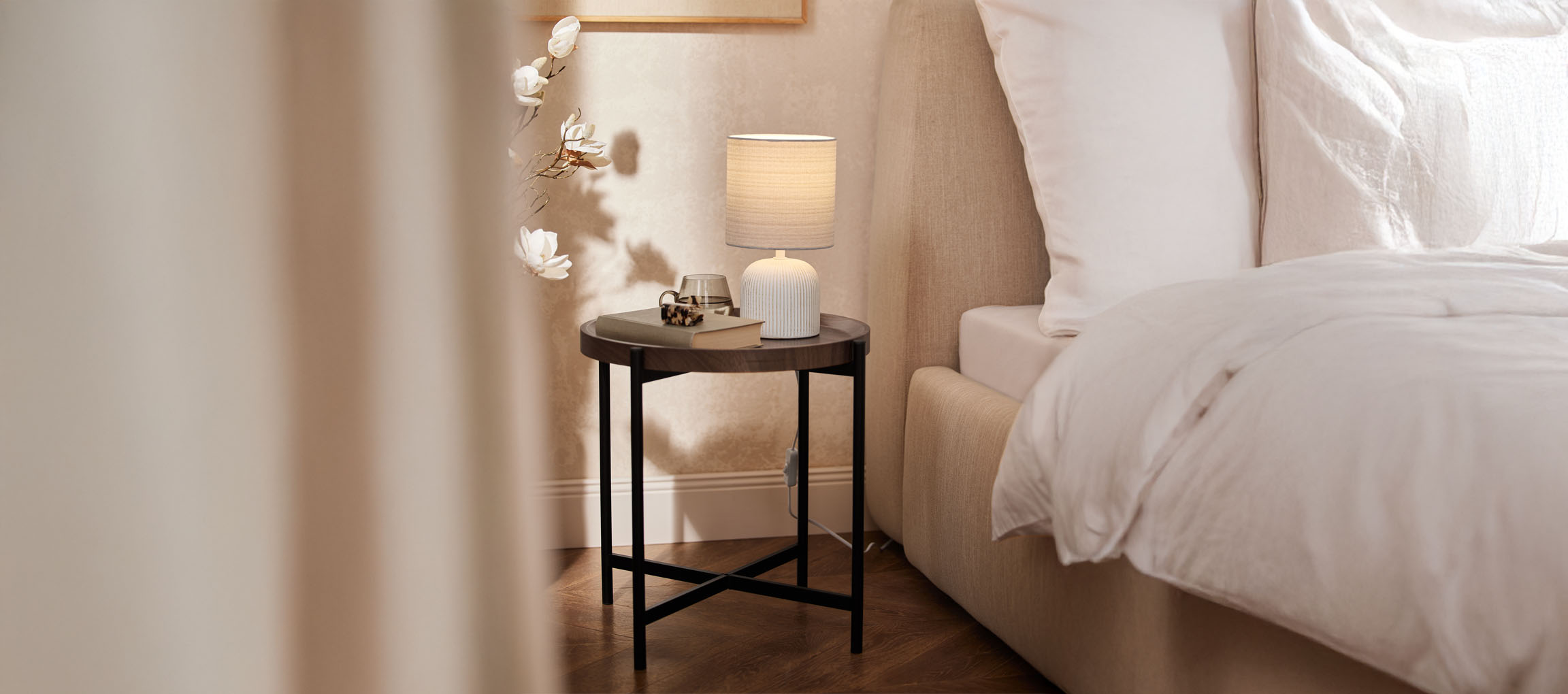 Runt sängbord i trä med bordslampa och beige säng i ett mysigt sovrum.