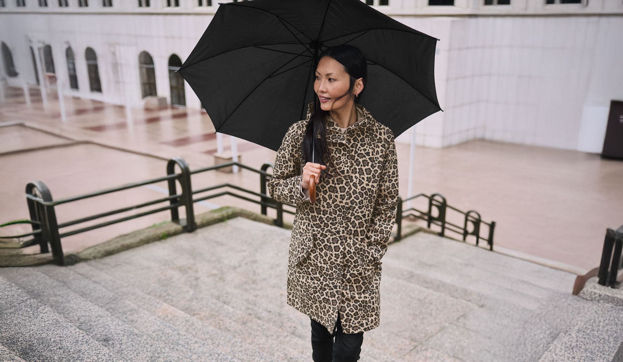 En kvinna i leopardmönstrad kappa håller ett svart paraply på en trappa.