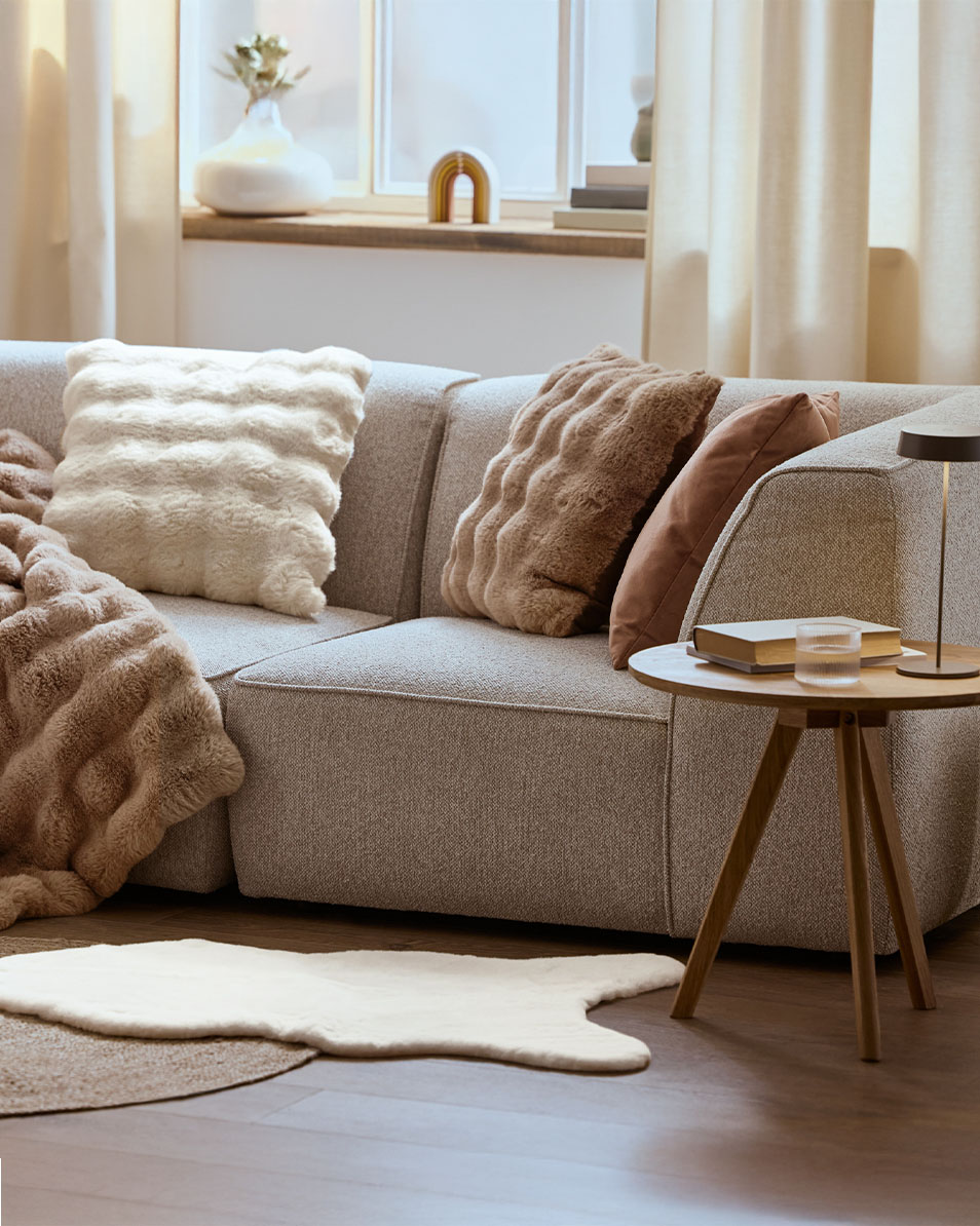 Grå modulsoffa med fluffiga kuddar och filtar, sidobord i trä.
