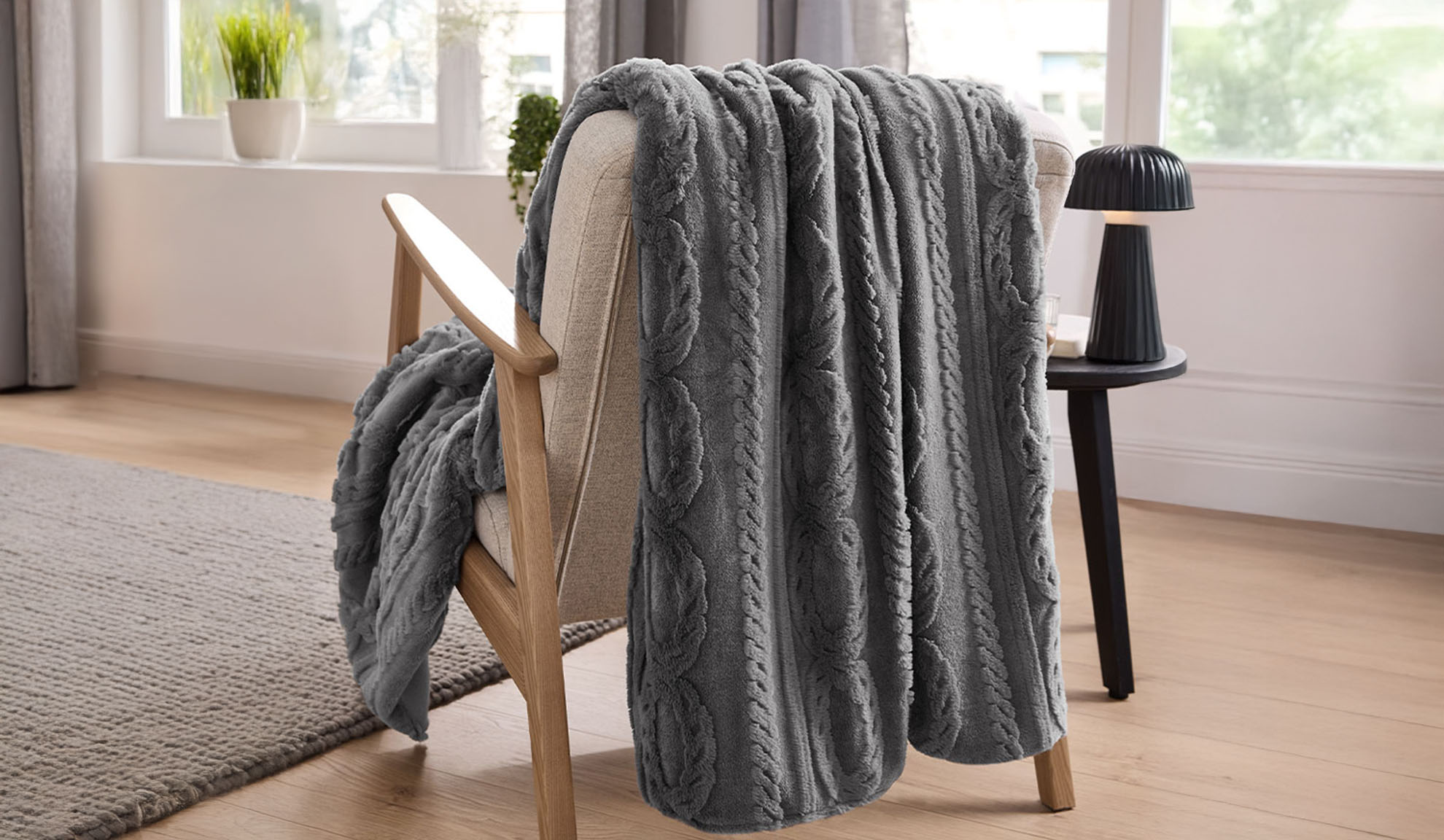 En grå kabelstickad pläd draperad över en ljus träfåtölj i ett vardagsrum.