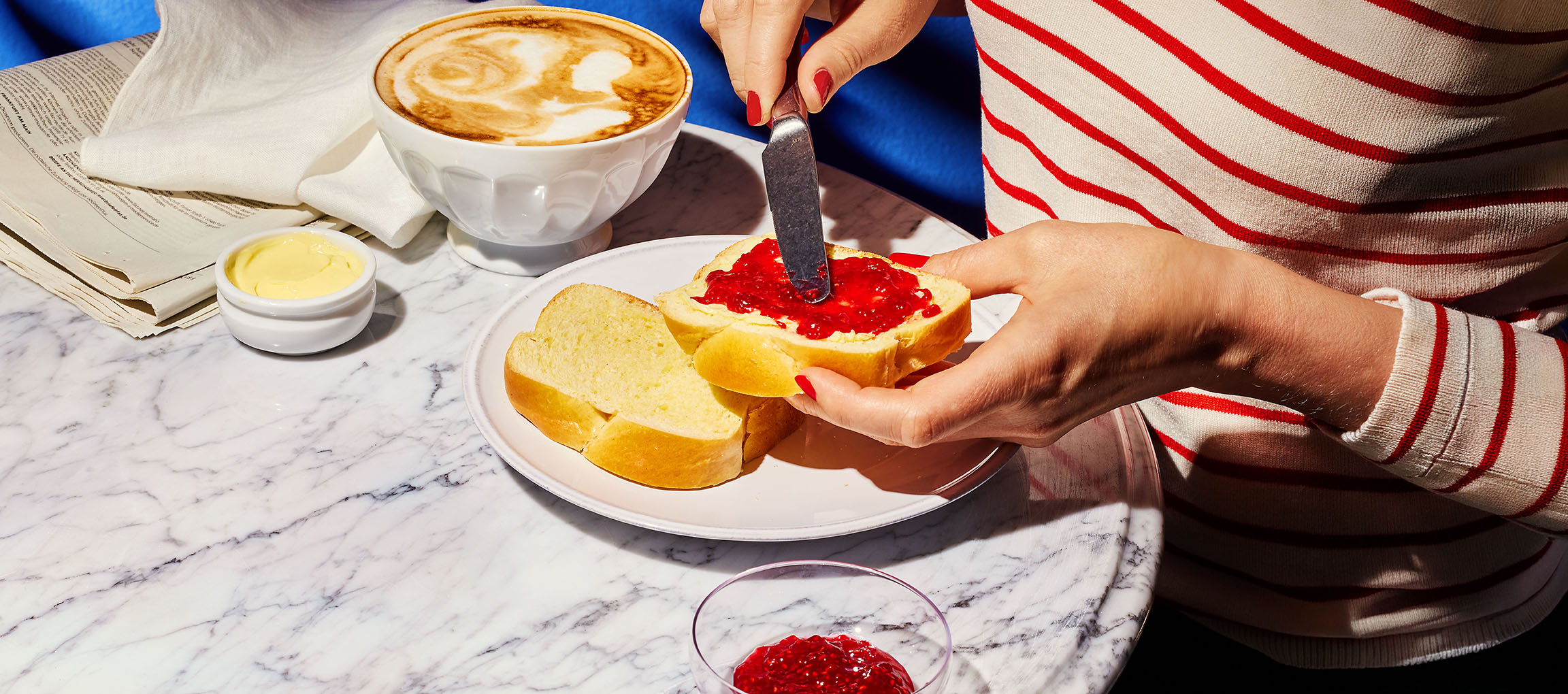 Händer brer sylt på briochebröd, med kaffe och smör på ett marmorbord.