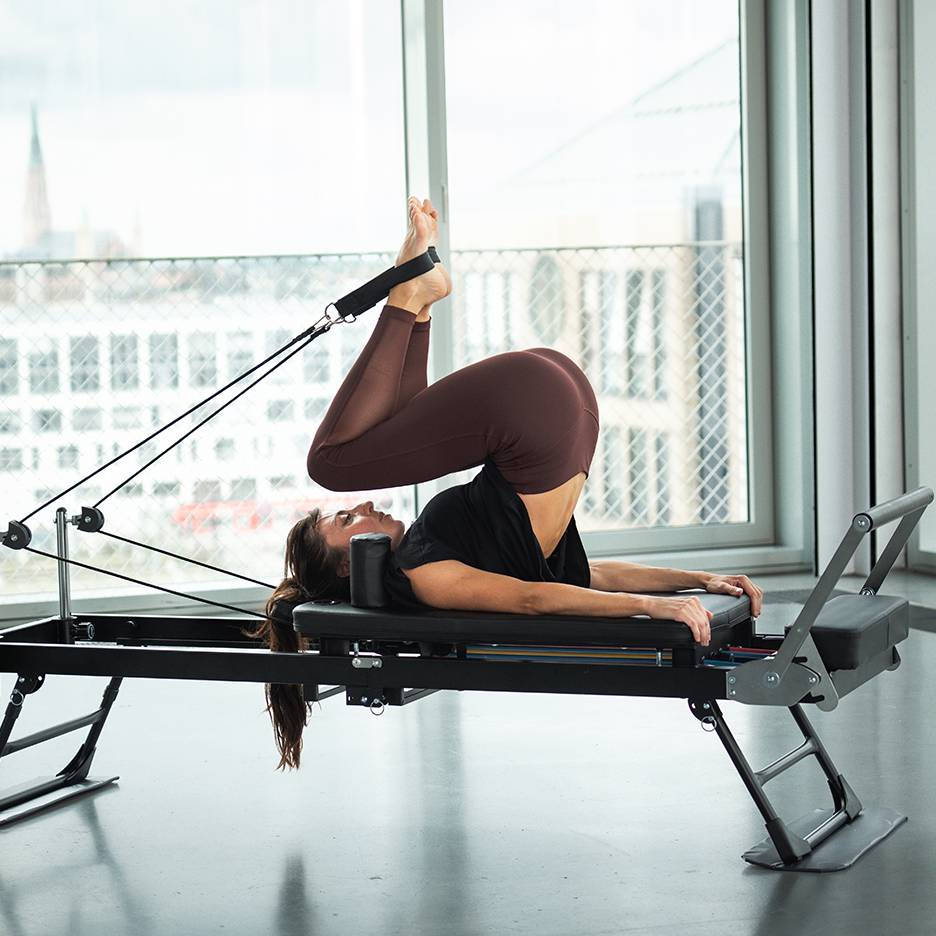 Kvinna tränar pilates på en reformer med benen uppåt och fötterna i remmar.