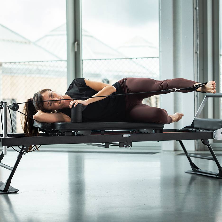 Kvinna tränar pilates på en reformer-maskin, liggande på sidan med benremmar.