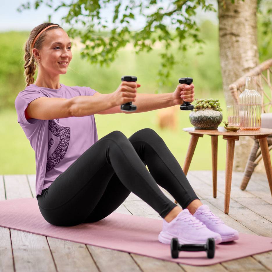 Kvinna i lila t-shirt och svarta leggings tränar med hantlar på en yogamatta.
