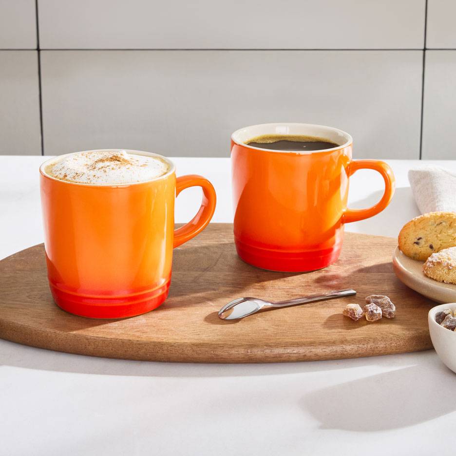 Två orange kaffemuggar på en träbricka, en med skummig kaffe, den andra med svart kaffe.