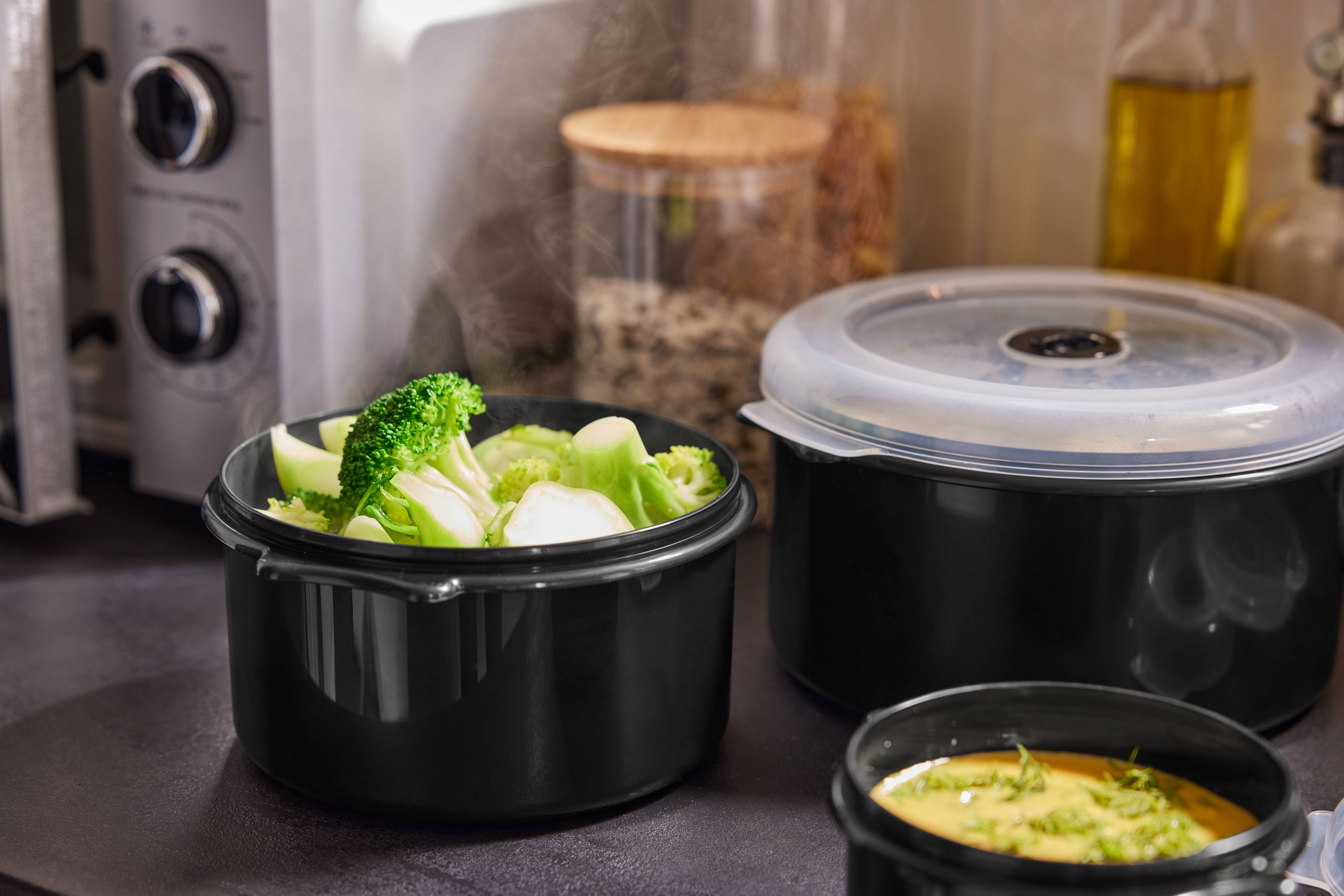 Ångande broccoli och andra rätter i svarta behållare på köksbänken.