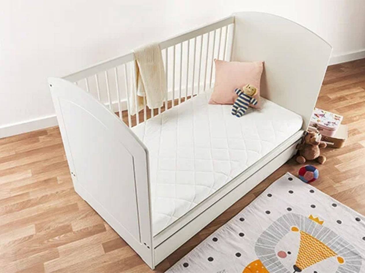 Vit babysäng med madrass, kudde och leksaker på ett trägolv.