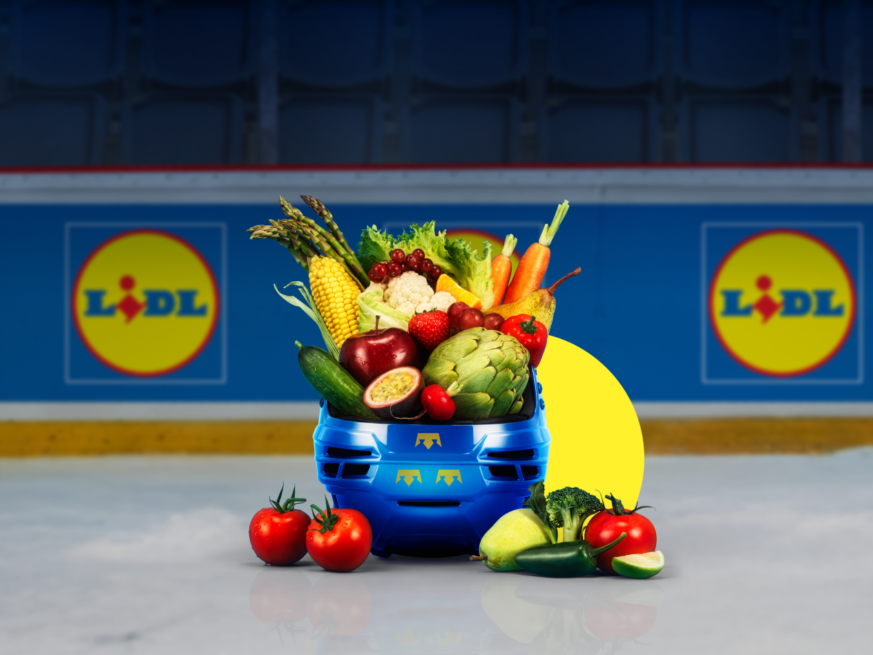 En blå hockeyhjälm fylld med färsk frukt och grönsaker på en isrink med reklamskyltar i bakgrunden.