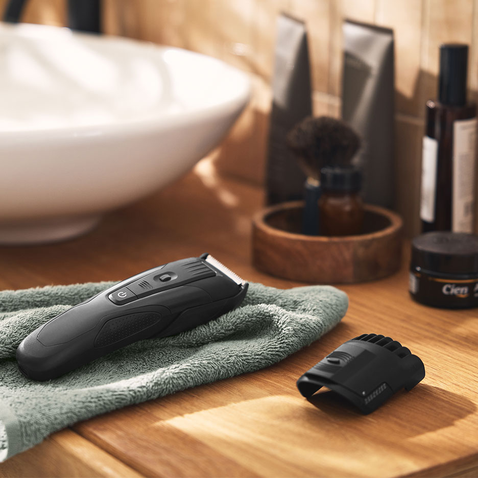 Svart hårtrimmer på en grön handduk i ett badrum med handfat och groomingprodukter.