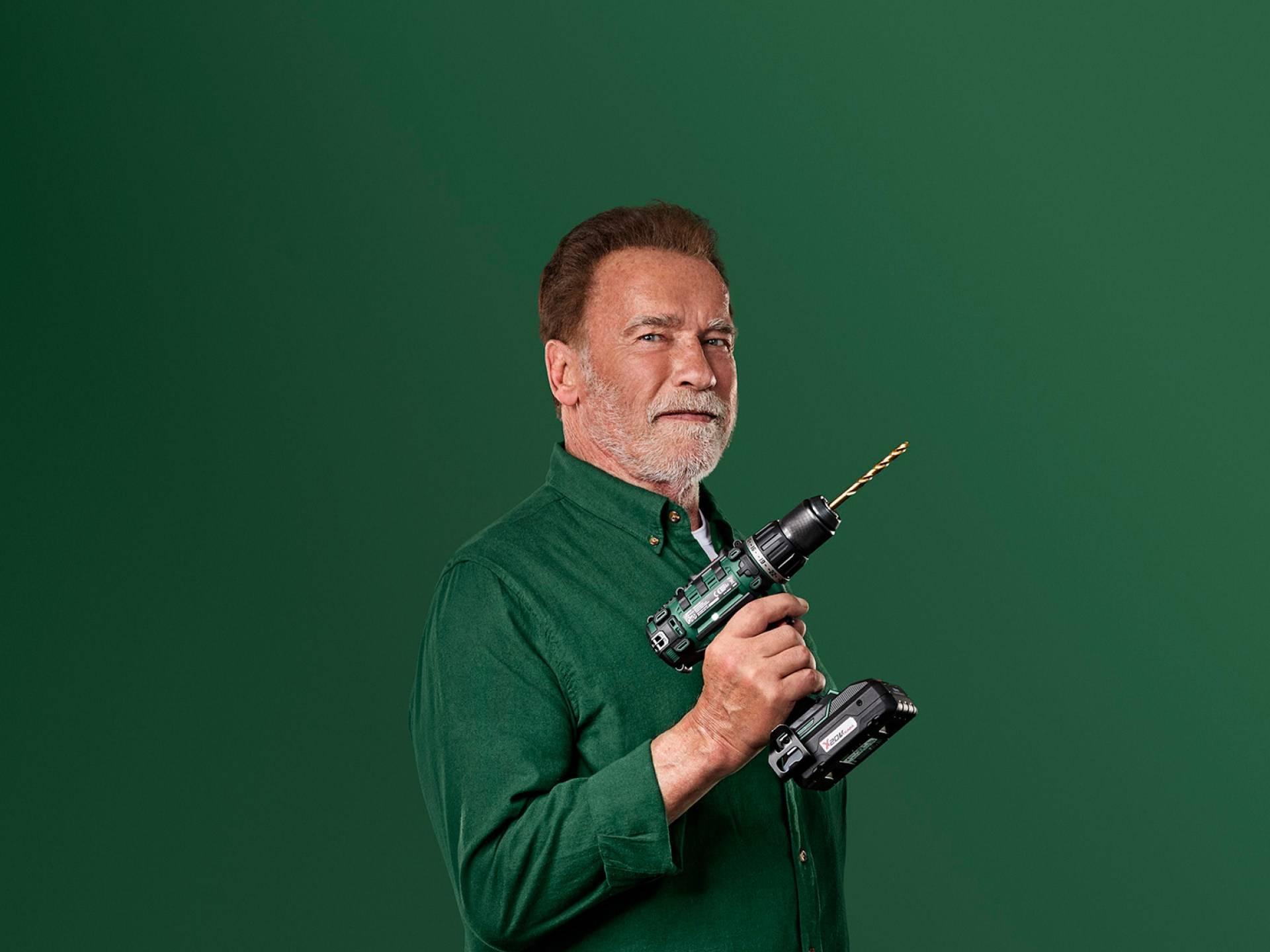 Arnold Schwarzenegger håller en grön elektrisk borrmaskin mot en grön bakgrund.