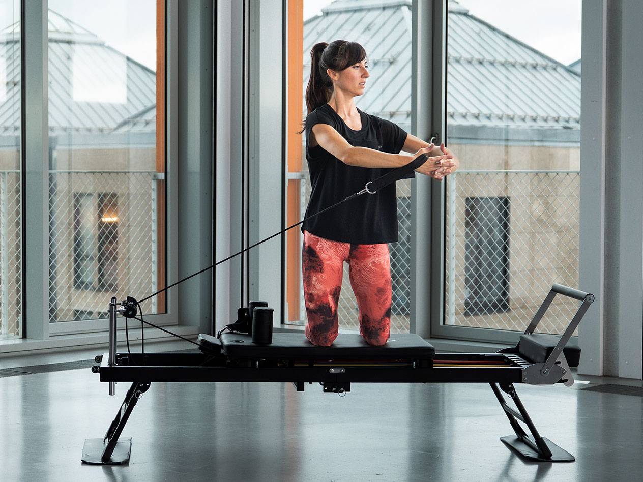 Kvinna tränar pilates på en svart reformer-maskin i ett ljust rum.