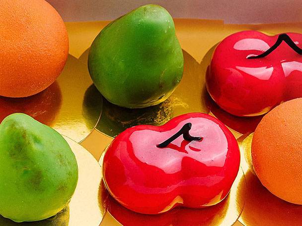 Färgglada fruktformade bakverk, inklusive röda äpplen, gröna päron och apelsiner, på en guldbakgrund.