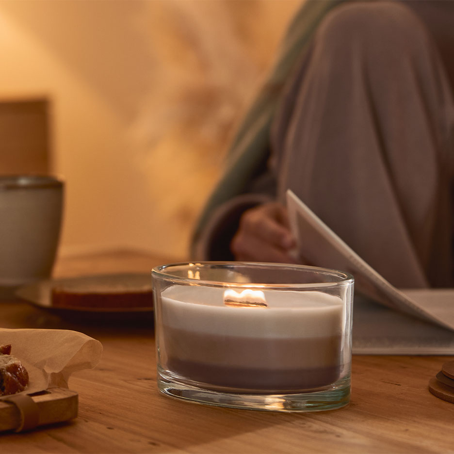 Brinnande trelagersljus i ett glas, med en person som läser i bakgrunden.