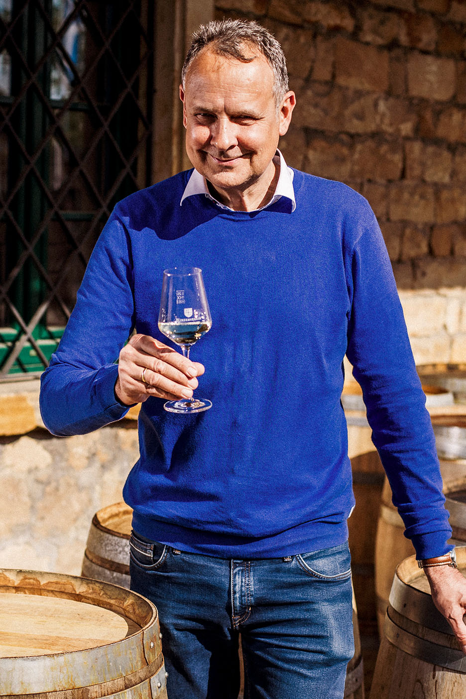 Leende man i blå tröja och jeans, håller ett glas vitt vin bland träfat.