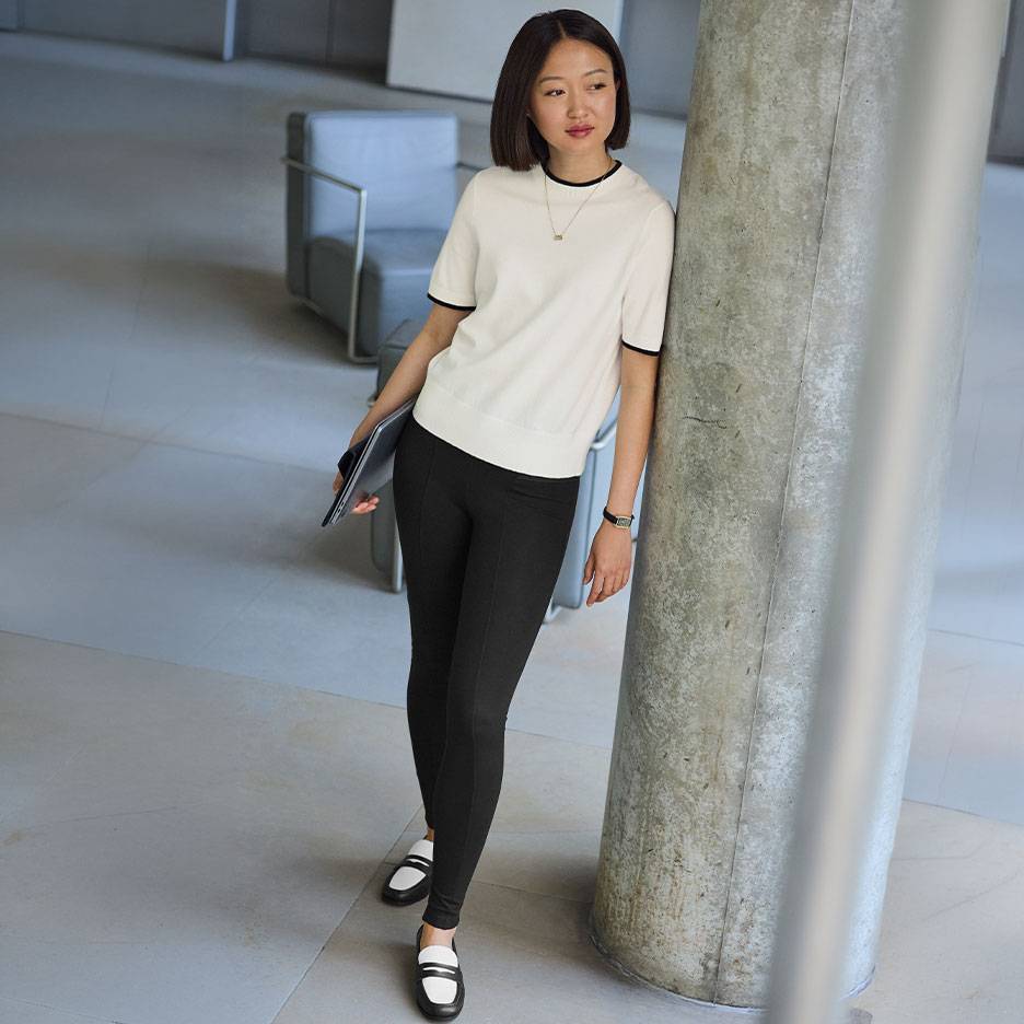 Kvinna i vit kortärmad topp med svart kant, svarta leggings och tvåfärgade loafers.
