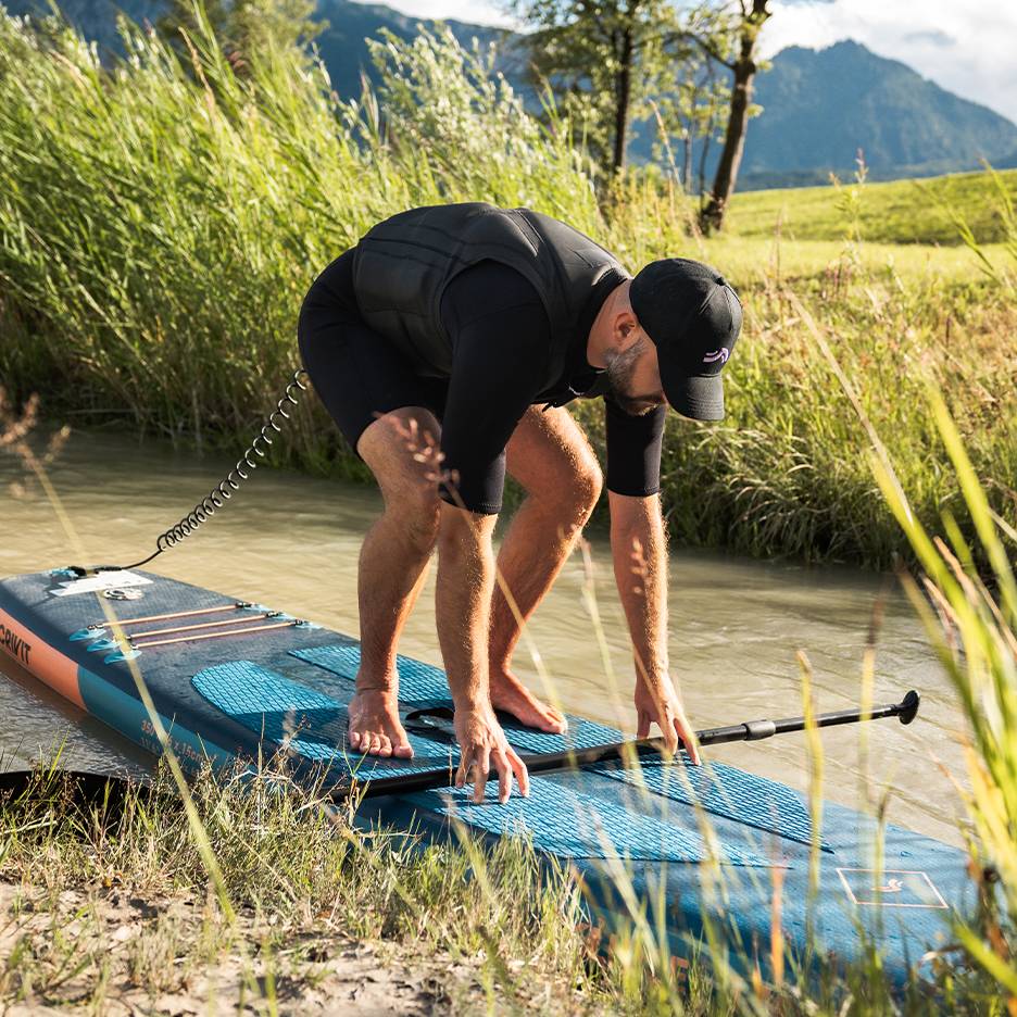 Man i våtdräkt och flytväst förbereder sig för stand-up paddling på en flod.