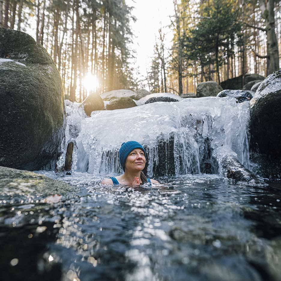 Kvinna i blå mössa och baddräkt njuter av ett kallbad i en frusen flod.