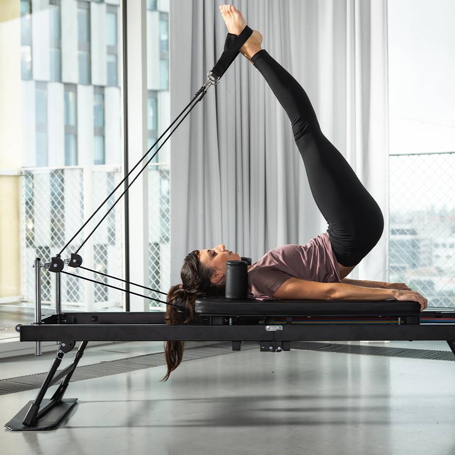 Kvinna tränar pilates på en svart pilatesmaskin med benen uppåt.