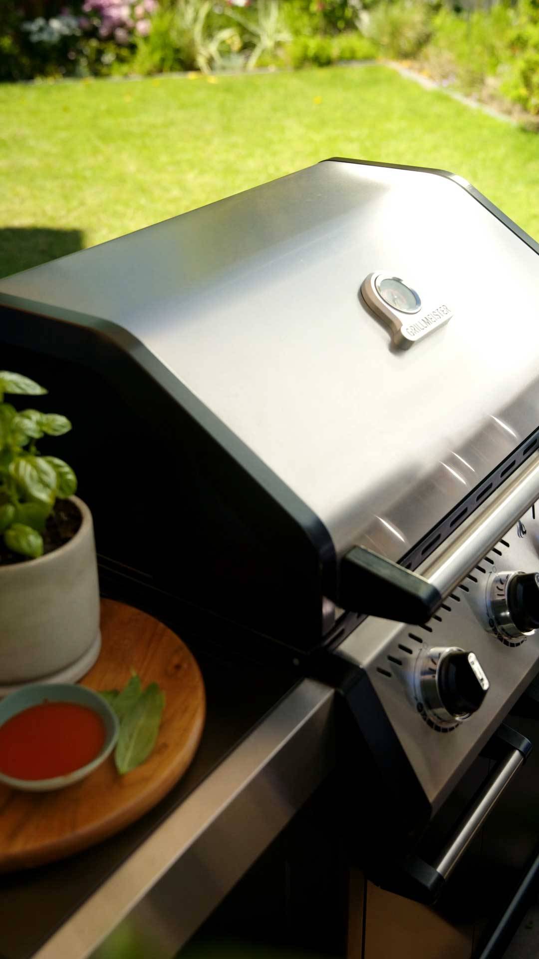 Gasolgrill i rostfritt stål med termometer, bredvid en basilika och sås.