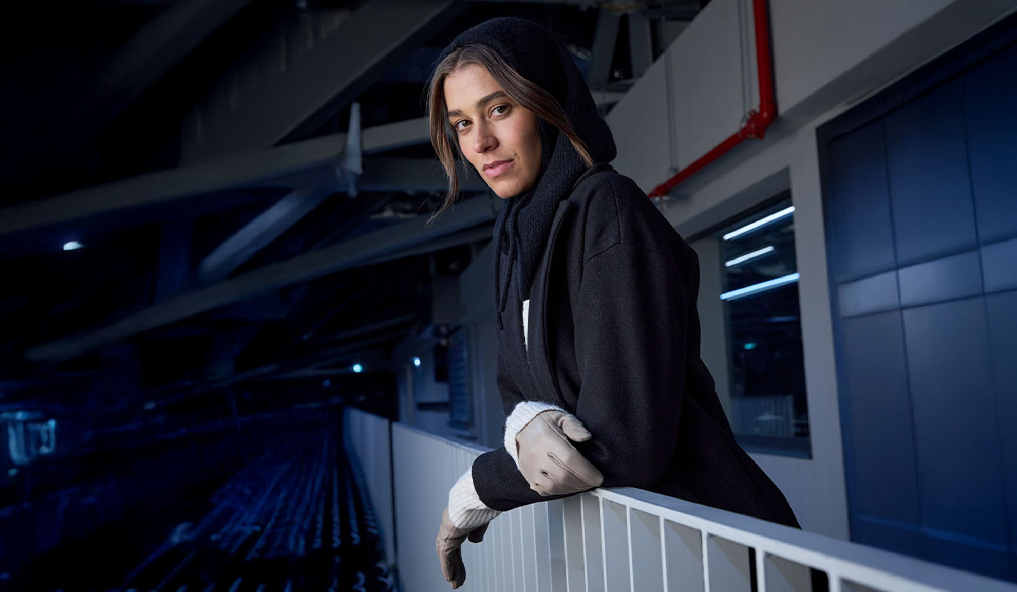 Kvinna i svart kappa och huvudsjal, lutad mot ett räcke i en modern miljö.