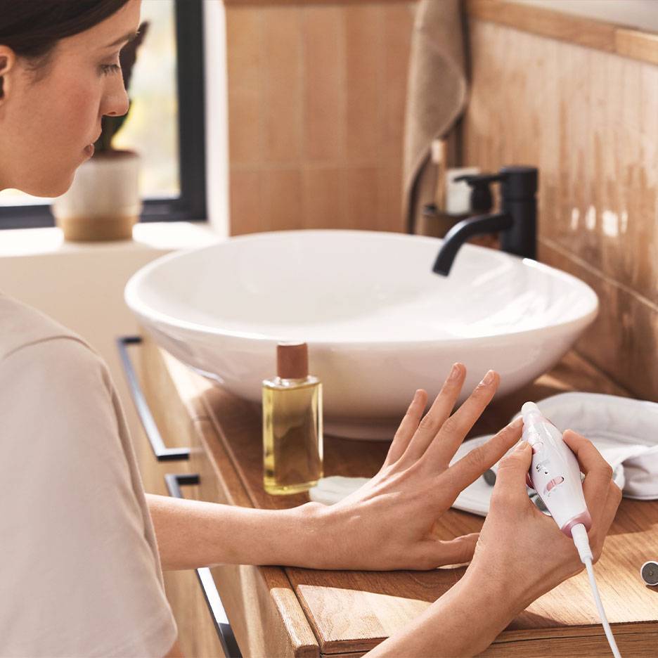 Kvinna använder ett elektriskt manikyr-/pedikyrset vid en träsminkbord med vitt handfat.