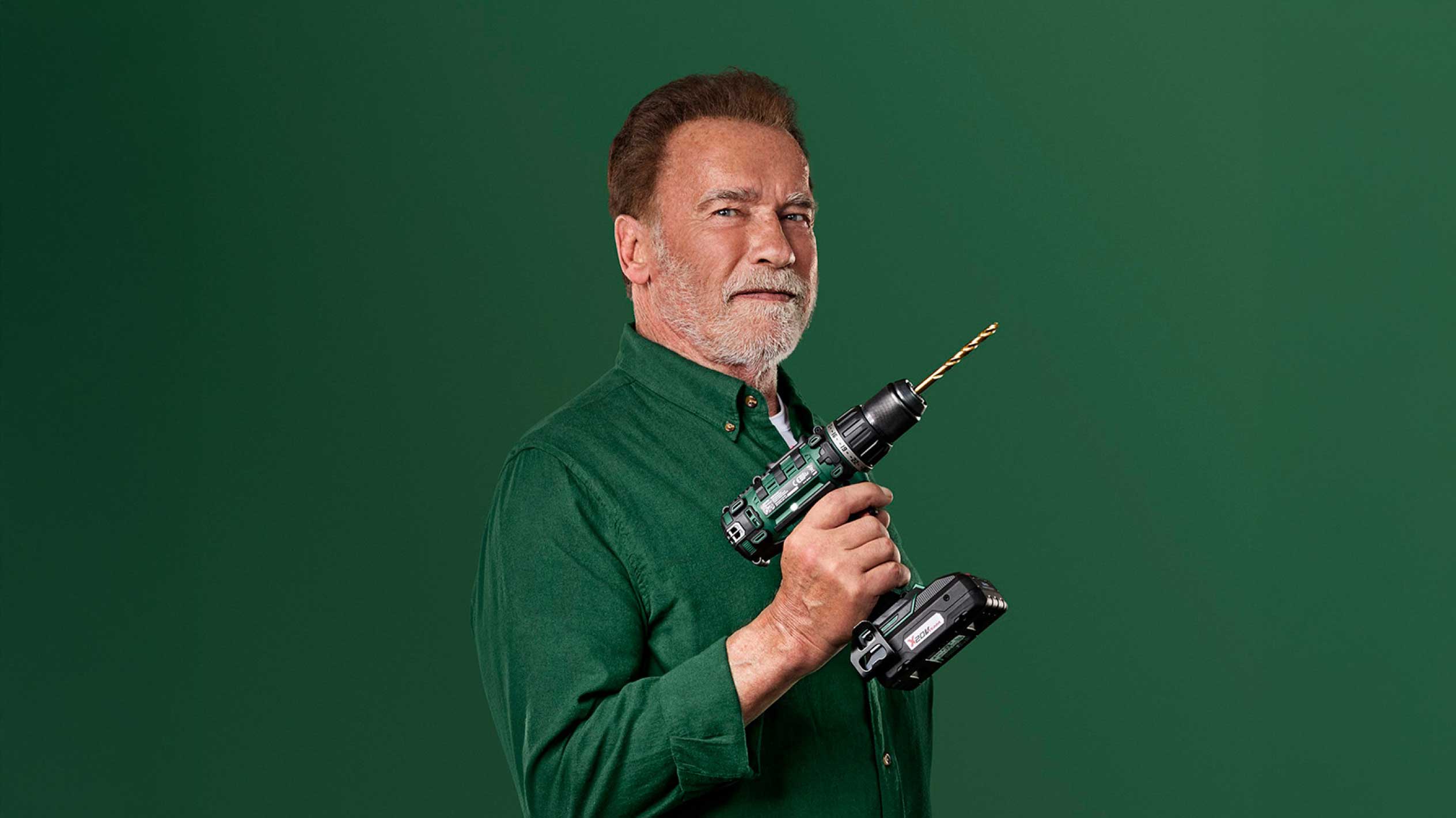 Arnold Schwarzenegger håller en grön sladdlös borrmaskin med ett guldfärgat borr.