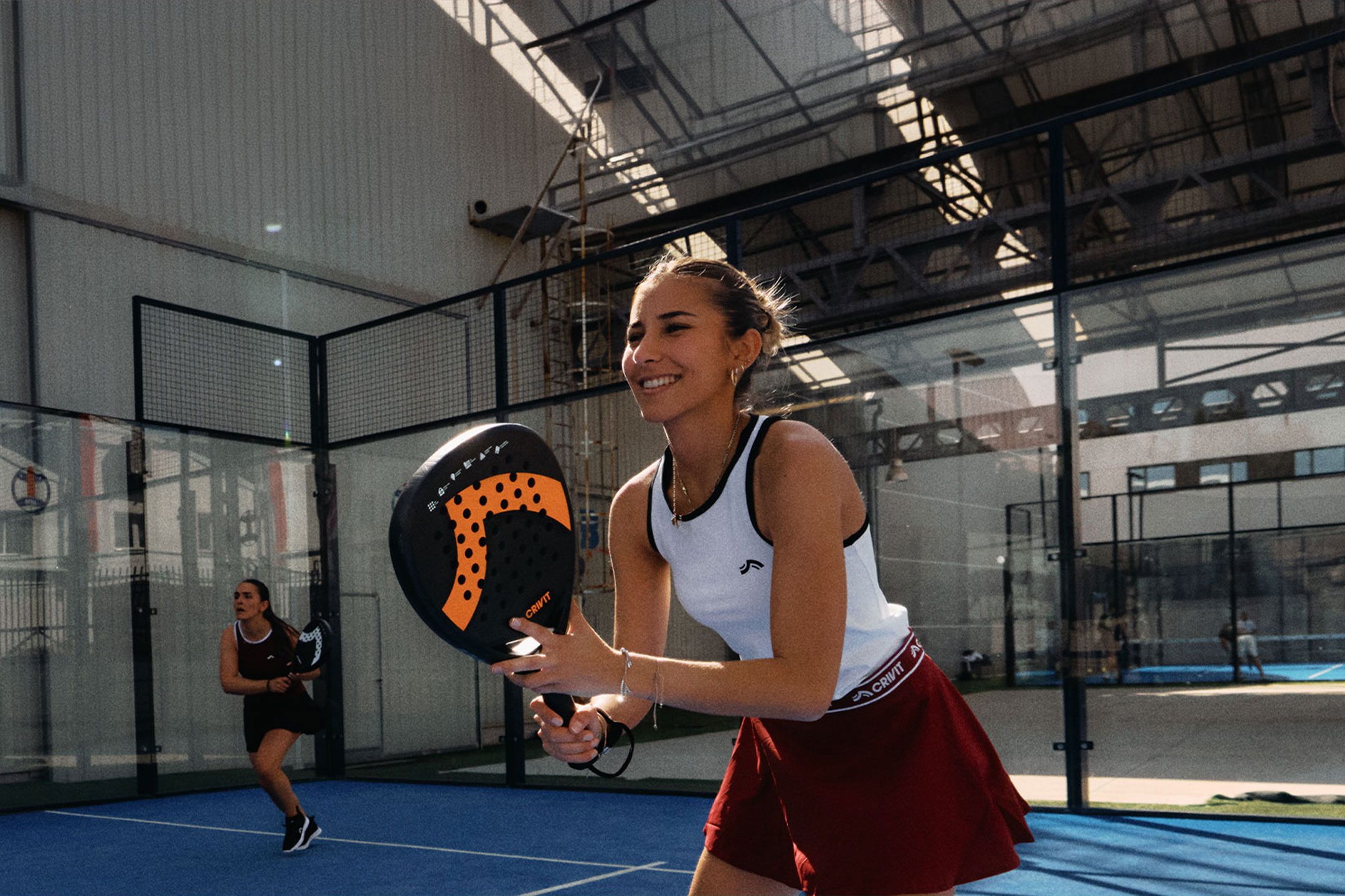 aiLabeling.aiLabelForImageAltText: Två kvinnor spelar padeltennis, en ler och håller ett racket, på en blå bana.