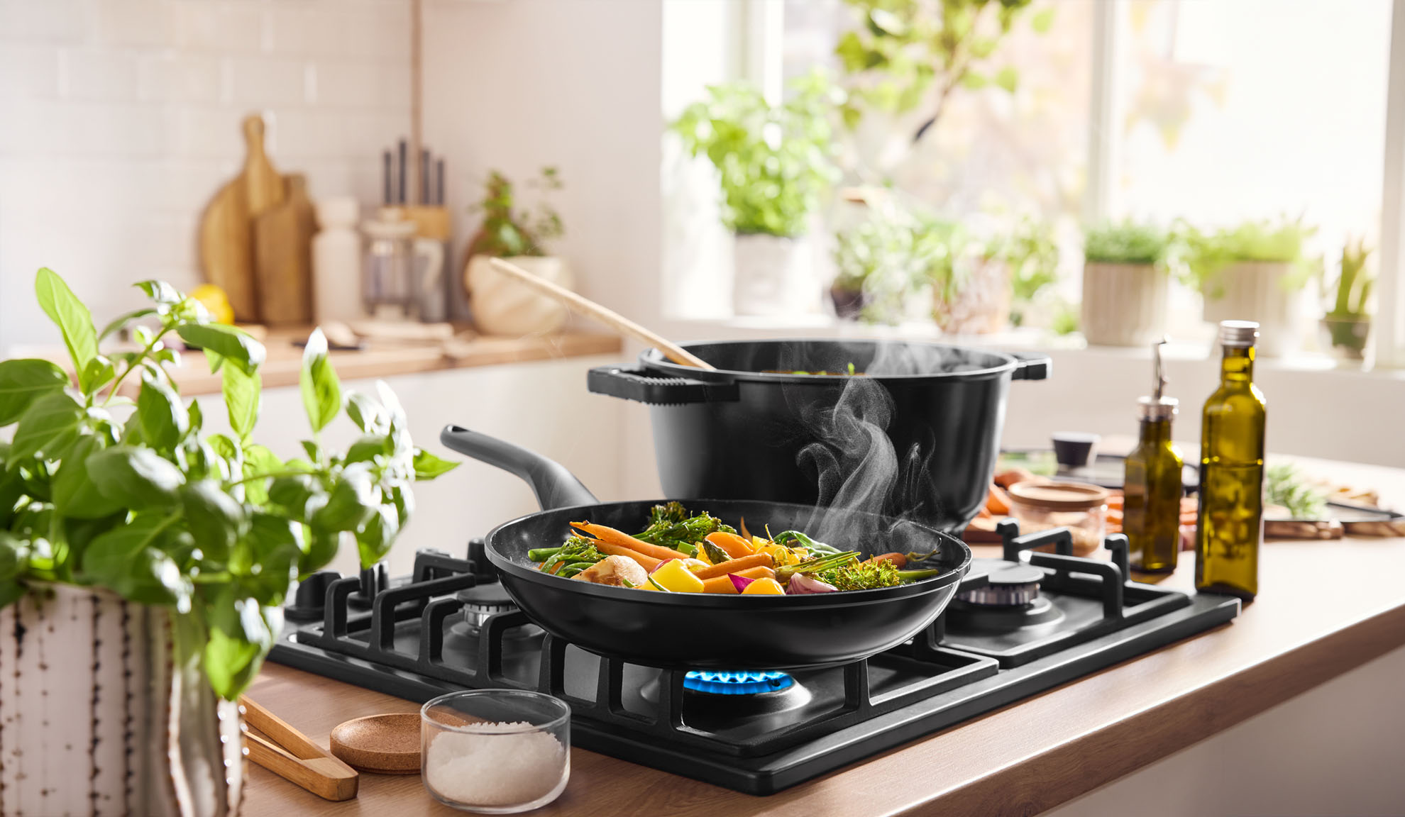 Ångande grönsaker kokar i panna och kastrull på gasspis i ett ljust kök.