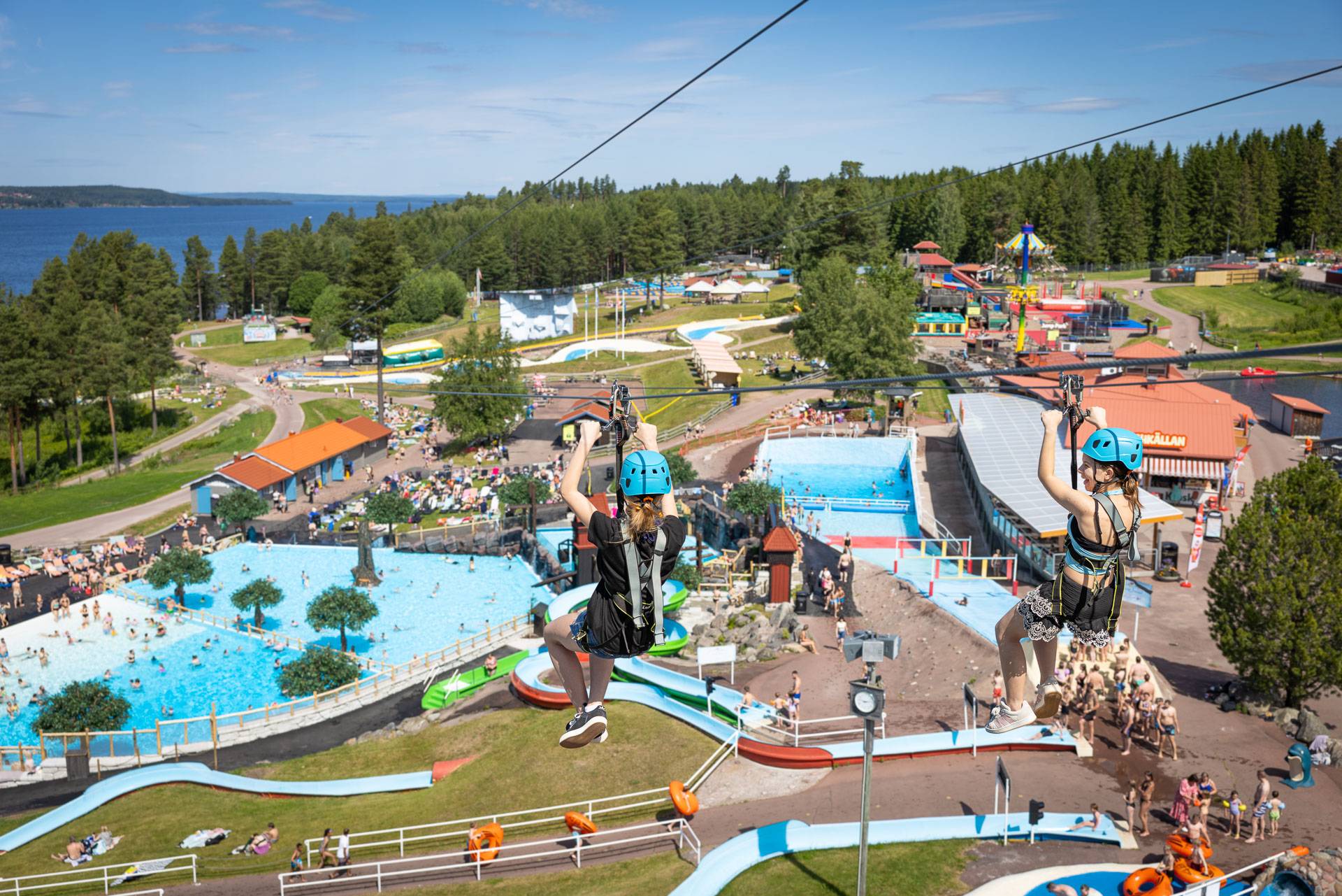 Två personer åker zipline över ett vattenland med pooler och vattenrutschbanor.