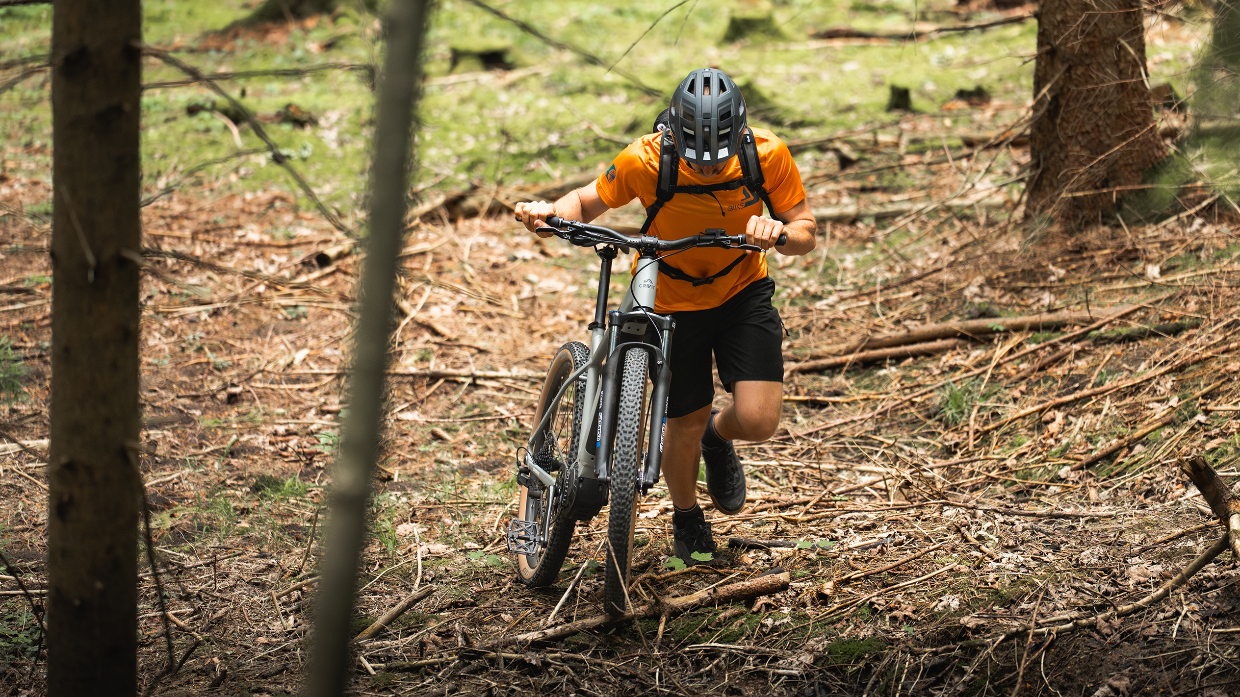 En cyklist i orange tröja puttar en eldriven mountainbike genom en skog.