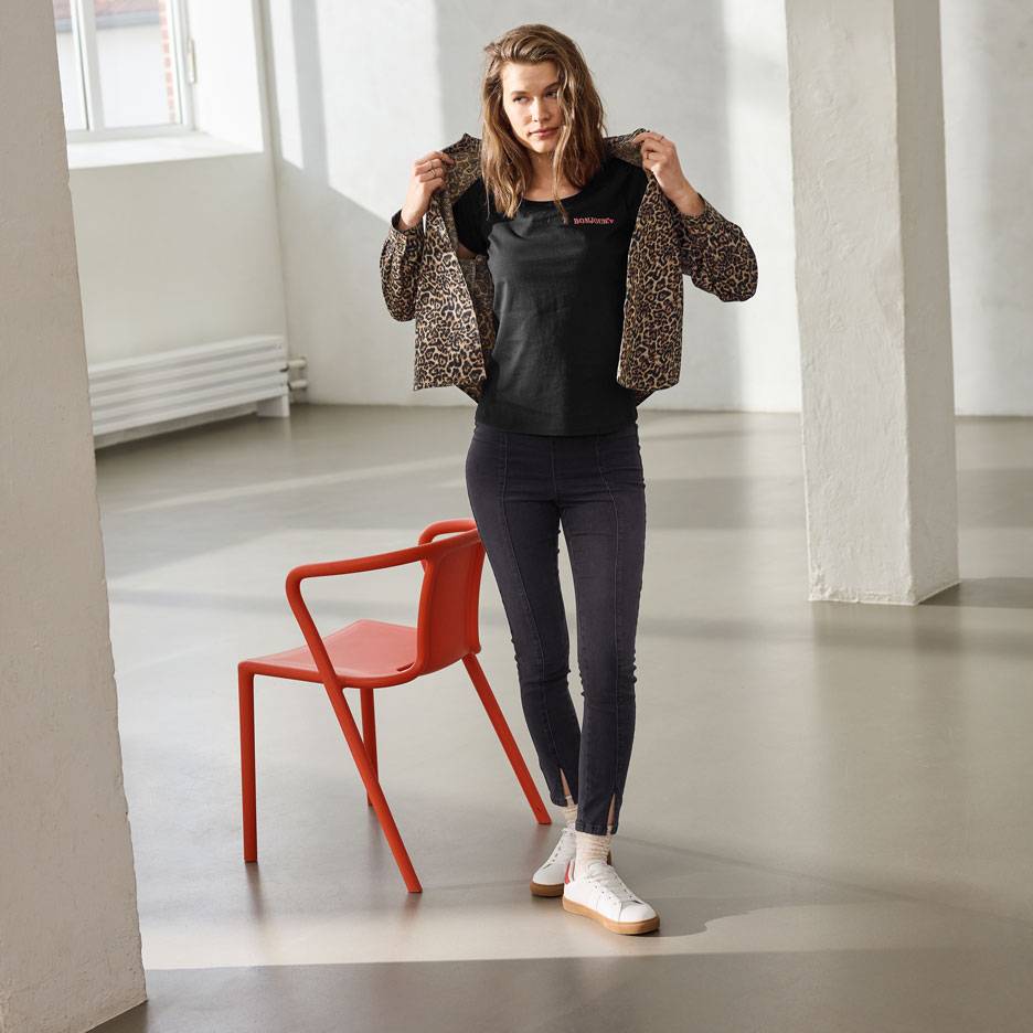 Kvinna i svart t-shirt med text, jeans och leopardmönstrad jacka, stående bredvid en röd stol.