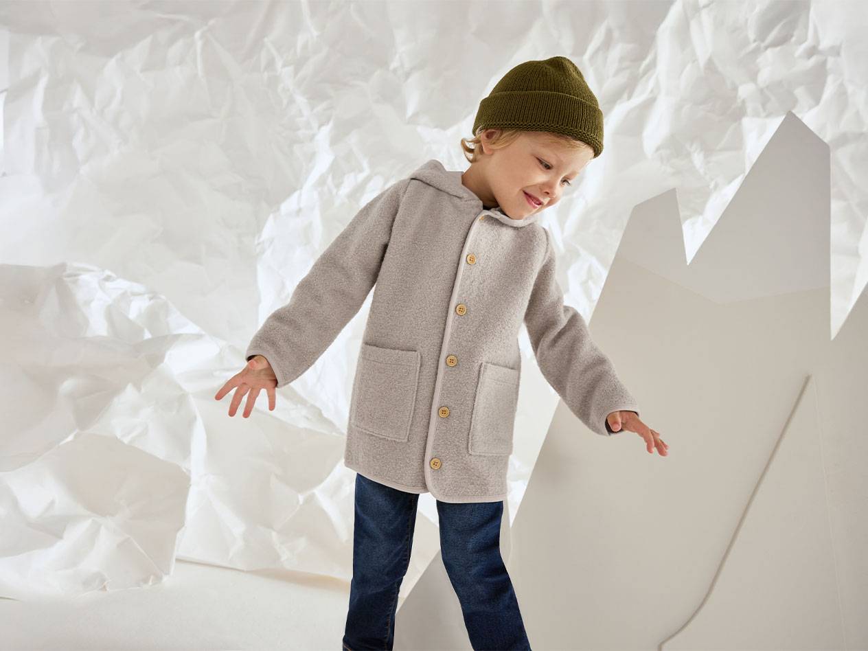 Pojke i grå yllejacka och grön mössa, leende mot en bakgrund av skrynkligt papper.