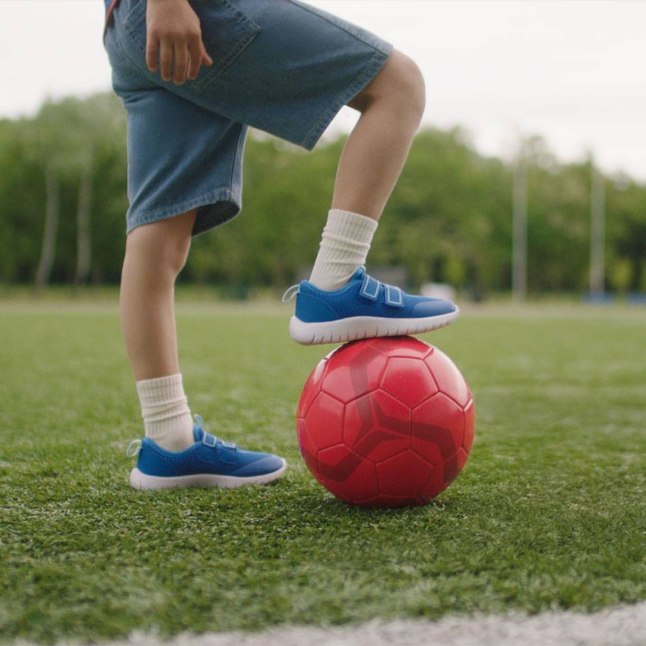 Barn i shorts och blå sneakers, med en fot på en röd fotboll.