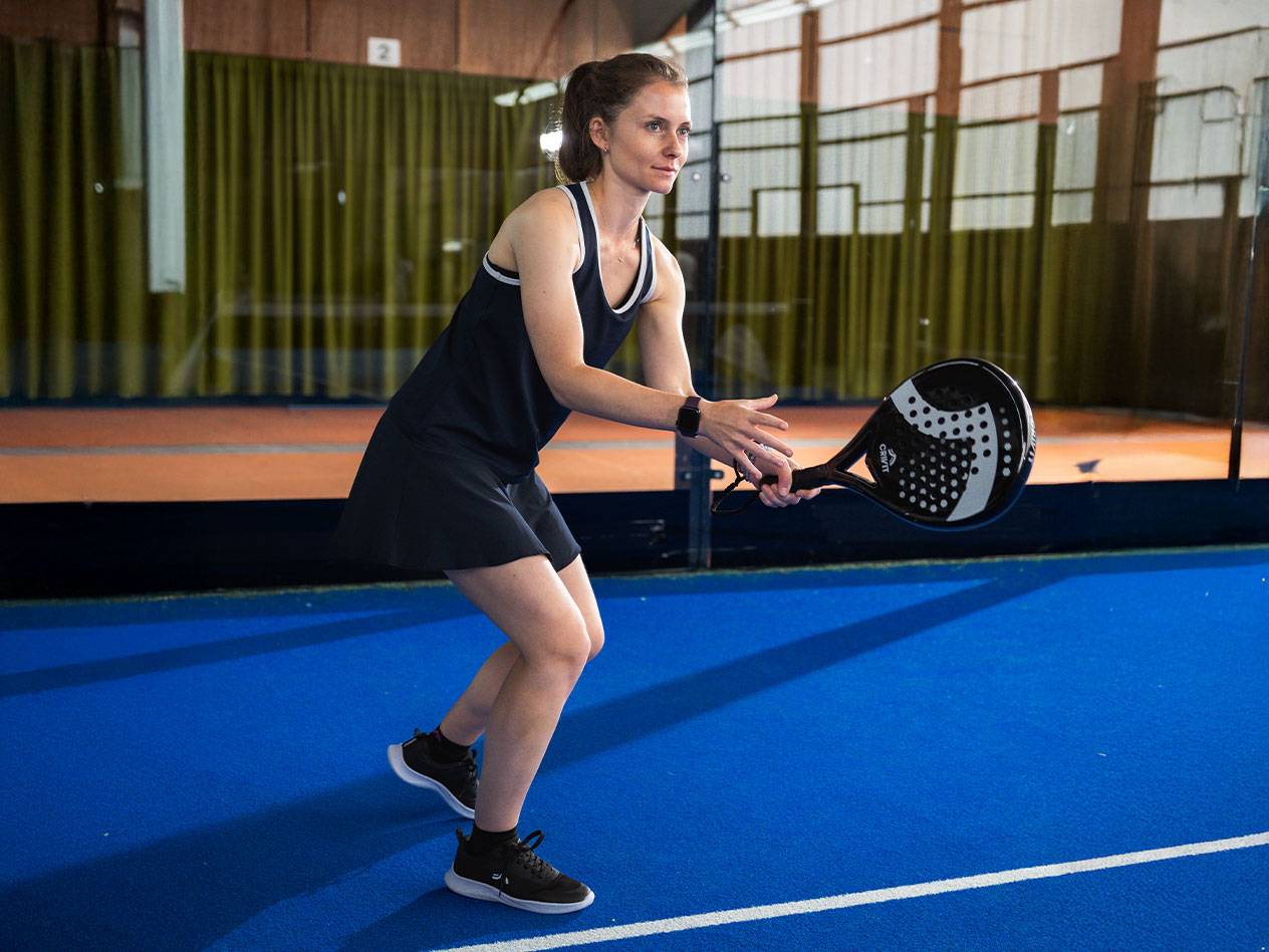 Kvinna i svart padelklänning med svart padelracket på blå bana.