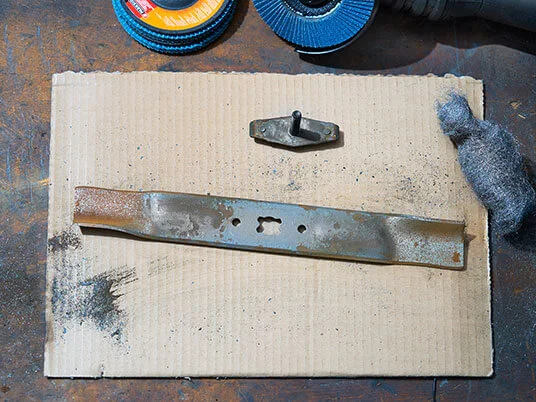 Rostigt gräsklipparblad på kartong med slipskivor och stålull.