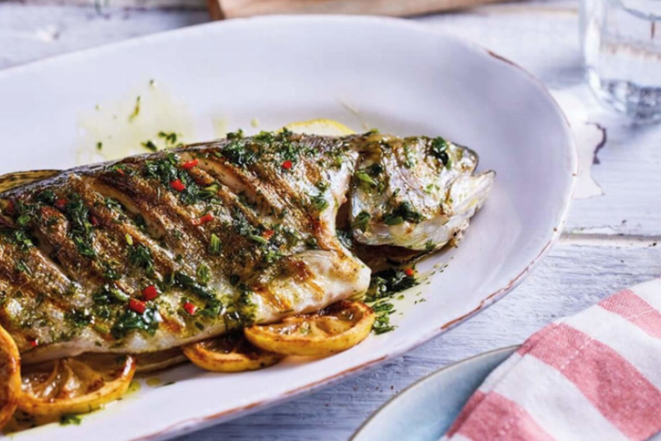 Grillad hel fisk med örter och citron på ett vitt fat.