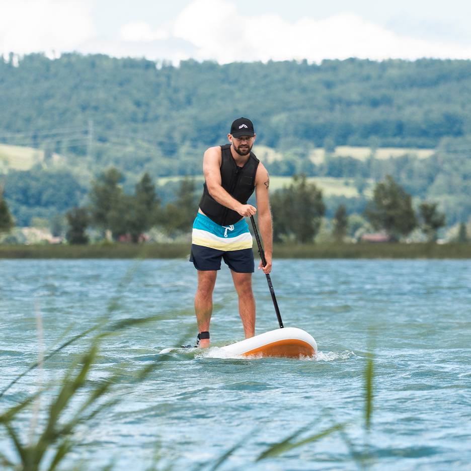 Man paddlar stand-up paddleboard på en sjö, med flytväst och badshorts.