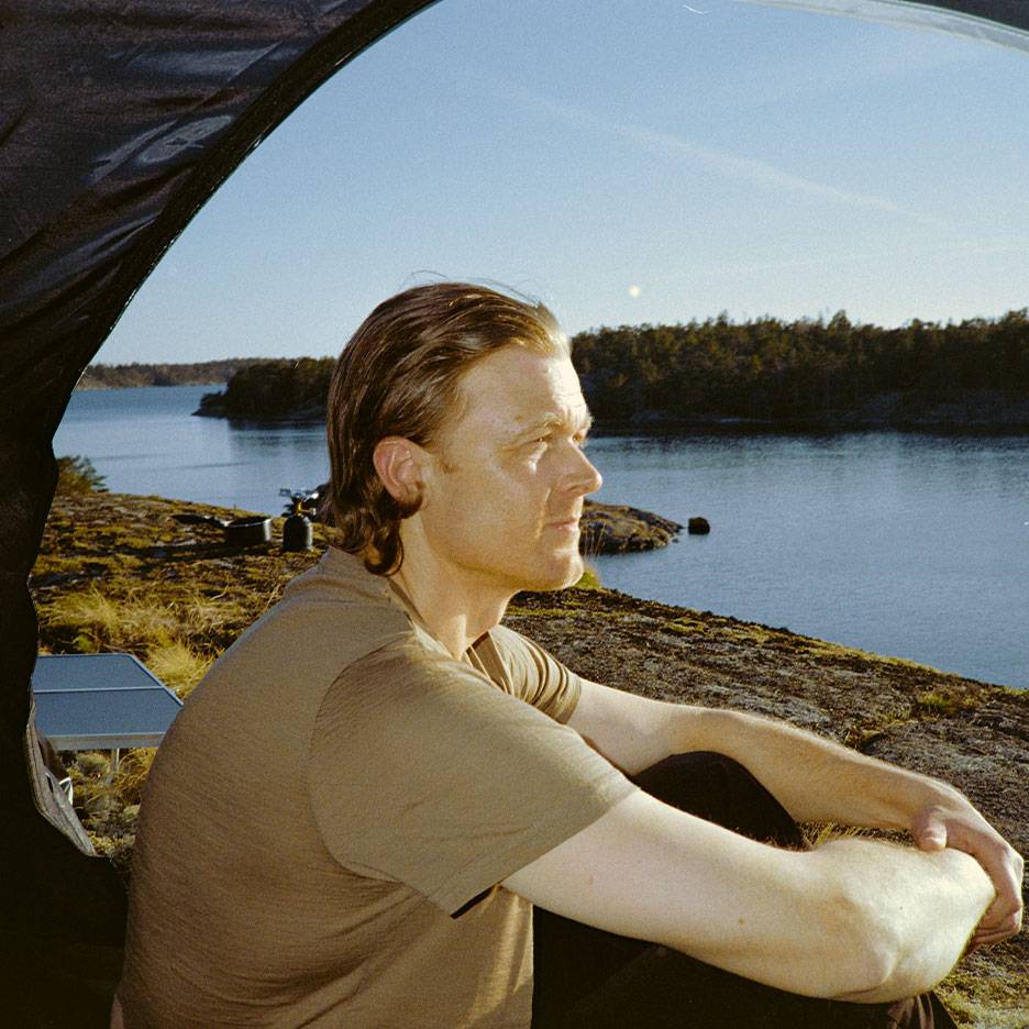 Man i sport-t-shirt sitter i tält, tittar ut över sjö och skog.